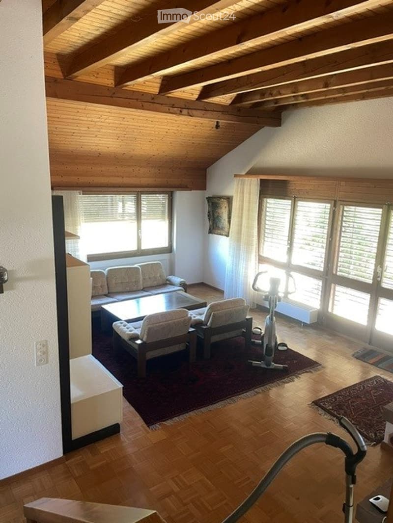 Bright living room with wooden beams, large windows, a sofa, coffee table, armchairs, and an exercise bike.