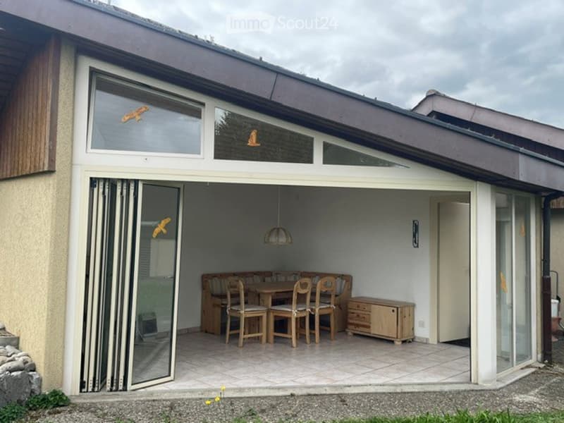 A covered outdoor terrace with large glass doors leading to the interior, featuring a dining table and chairs, a small cabinet, and a hanging light fixture.