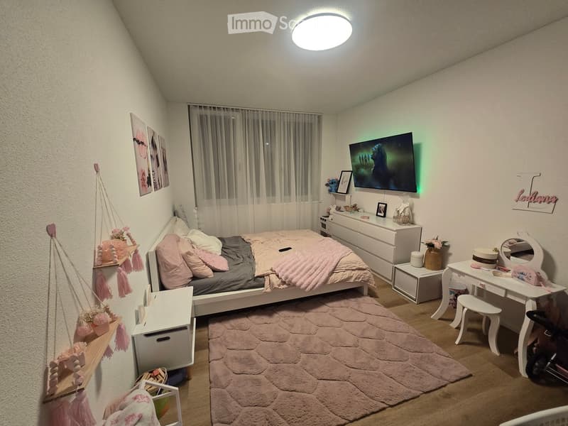Cozy bedroom with a bed, nightstand, mirror, stool, table, chair, and a large window with curtains. The room features a soft pink color scheme with decorative elements like a hanging basket and picture frames.