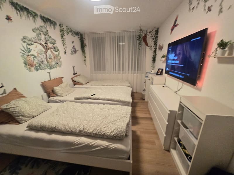 Cozy bedroom with two single beds, pillows, a nightstand, a television, and a curtained window. The walls feature a nature-themed mural with trees and animals.