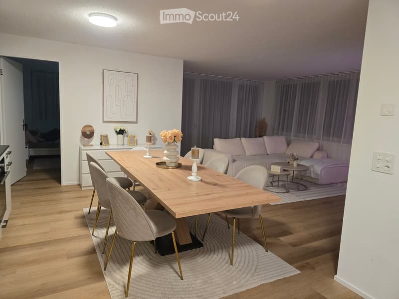 Modern living and dining area with a light wooden dining table, beige upholstered chairs, a grey sofa, side tables, decorative vases, and candles.