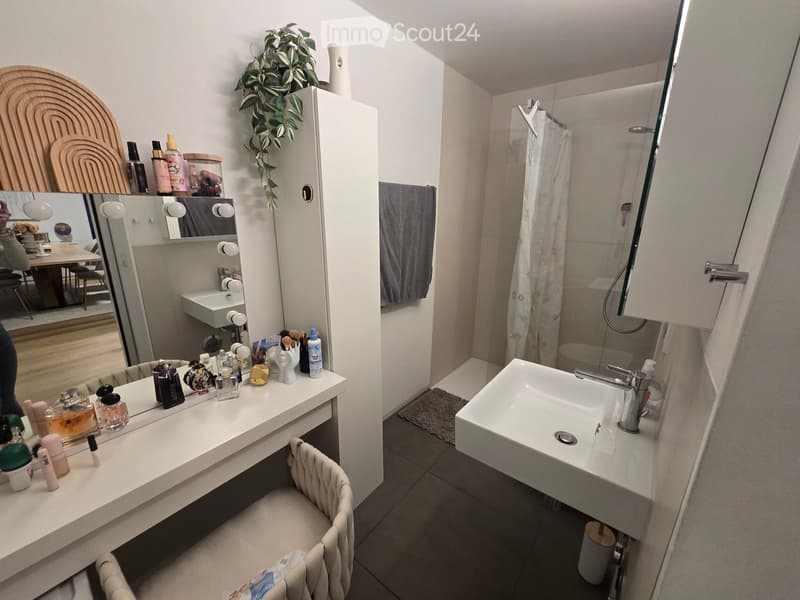 Modern bathroom with a white sink, large mirror, vanity with cosmetics, a small chair, and a shower area with a glass enclosure.