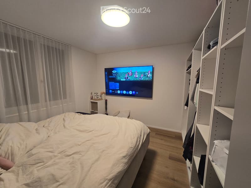 Modern bedroom with a large bed, mounted television, nightstand, and built-in wardrobe. The room features wooden flooring and large windows with curtains.
