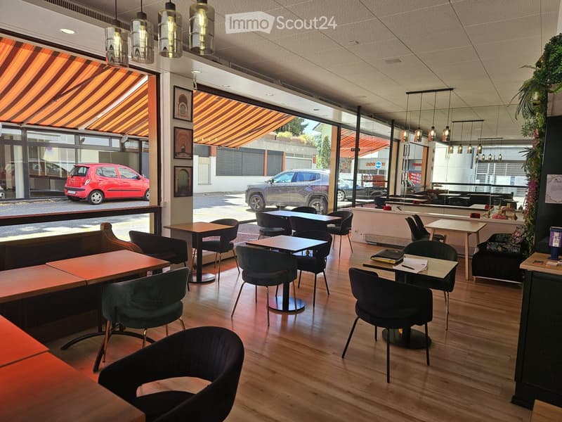 Modern restaurant interior with wooden tables, dark upholstered chairs, pendant lights, and framed artwork on the walls.