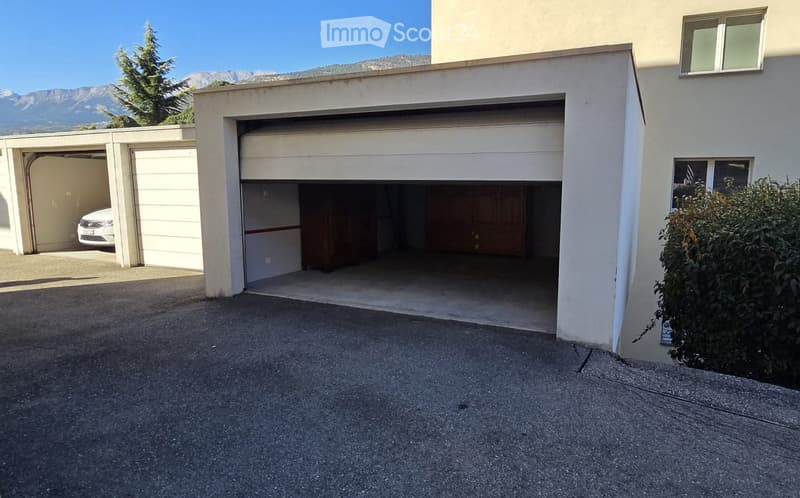 Two-car garage with a white car parked inside, located next to a building with a raised ground floor and a view of mountains in the background.