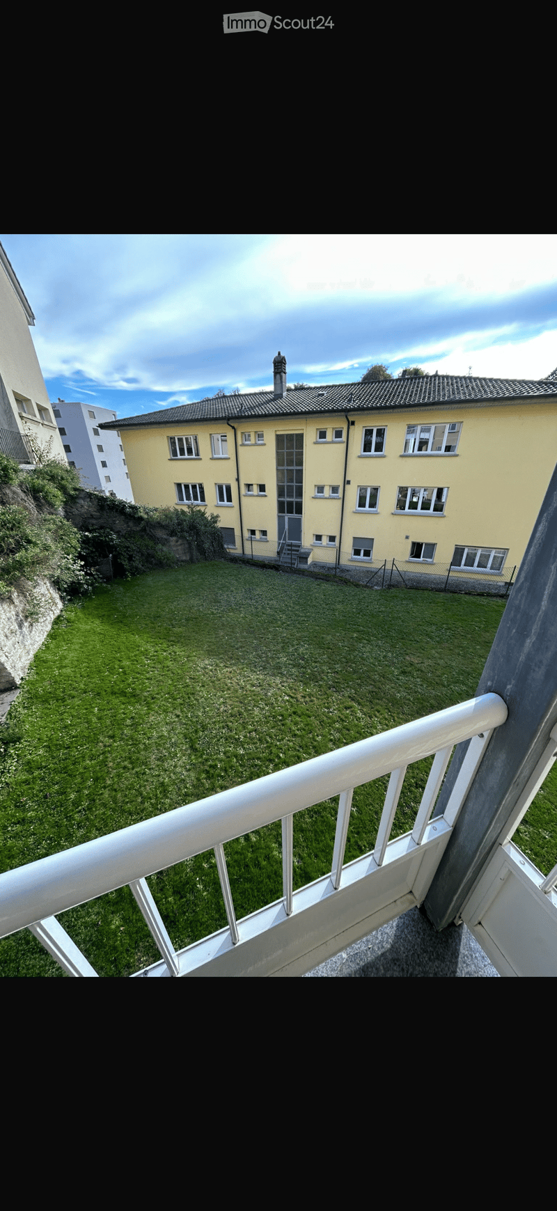 Balcony overlooking a grassy area with a yellow building in the background, featuring white railings and a view of the sky.