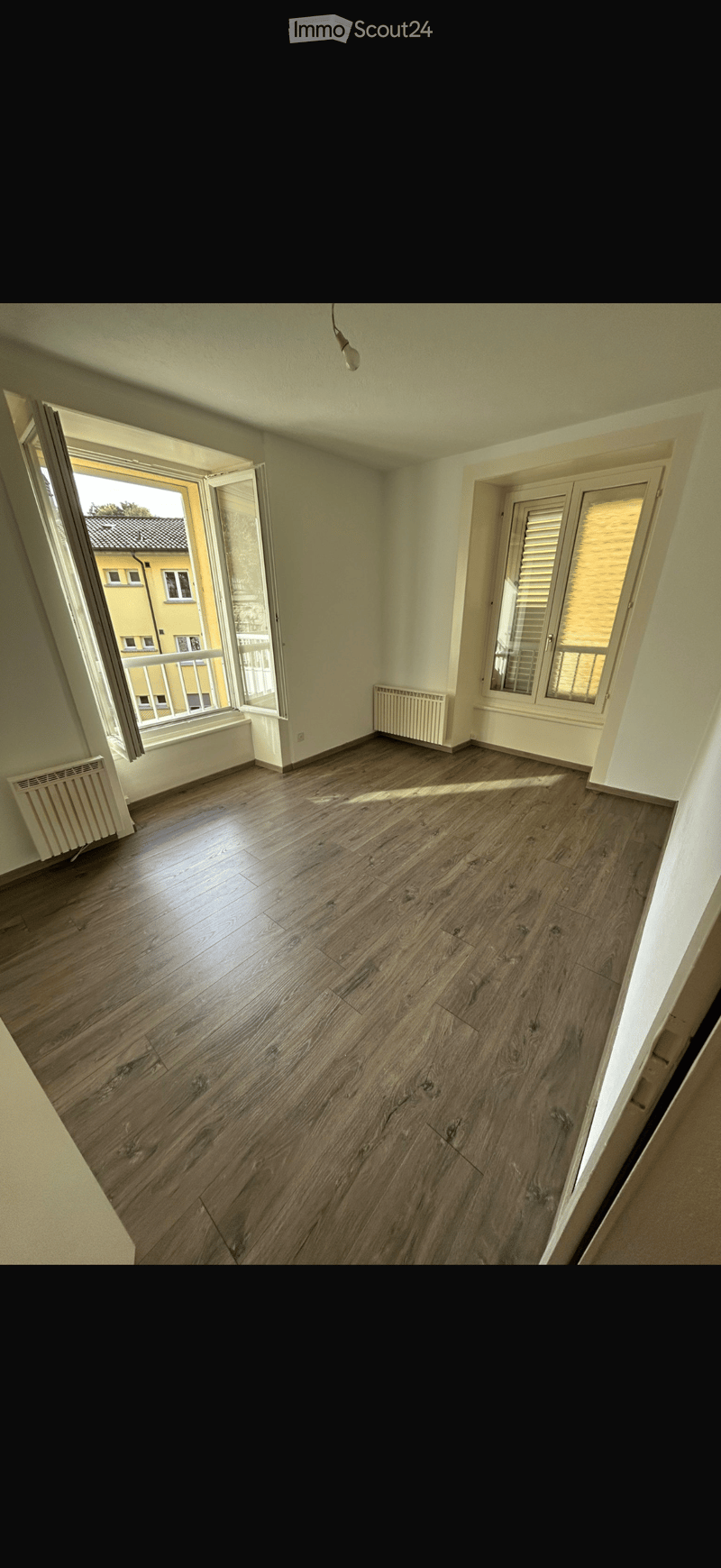 Empty room with wooden flooring, large windows, and radiators on the walls.
