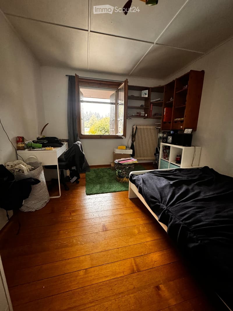 Cozy attic bedroom with wooden floors, slanted ceilings, and large window. Features a bed, desk, chair, bookshelf, radiator, and small green rug.