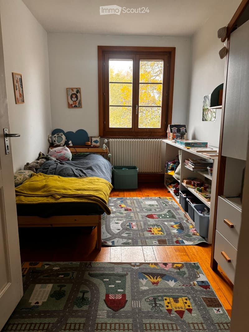 Cozy bedroom with wooden floors, large window, bed with yellow blanket, shelves with books and toys, and colorful rugs.