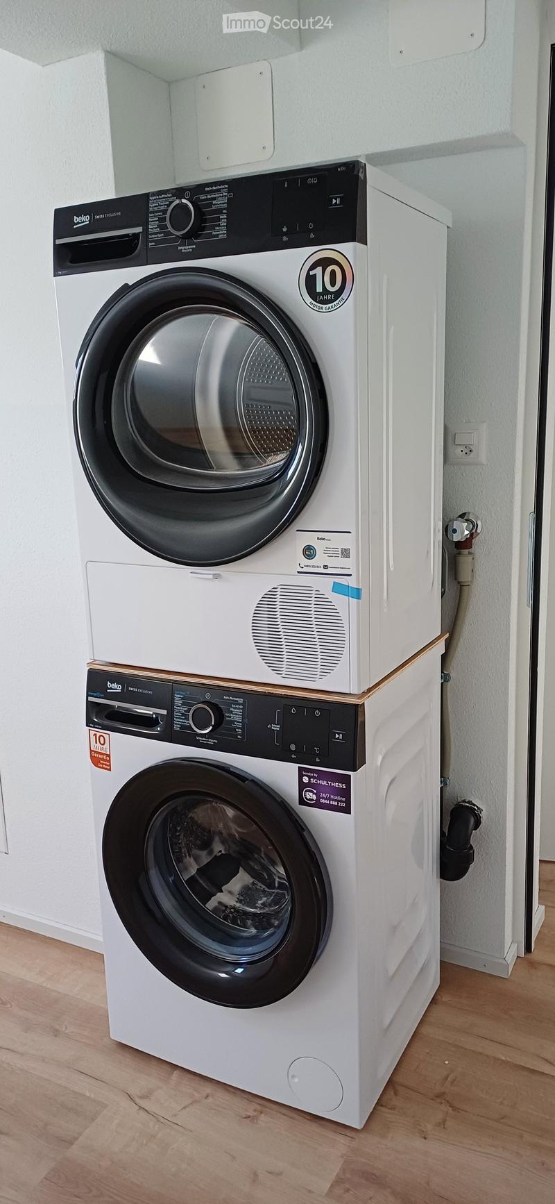 A modern, white washing machine and dryer unit is installed on a wooden floor in a bathroom.