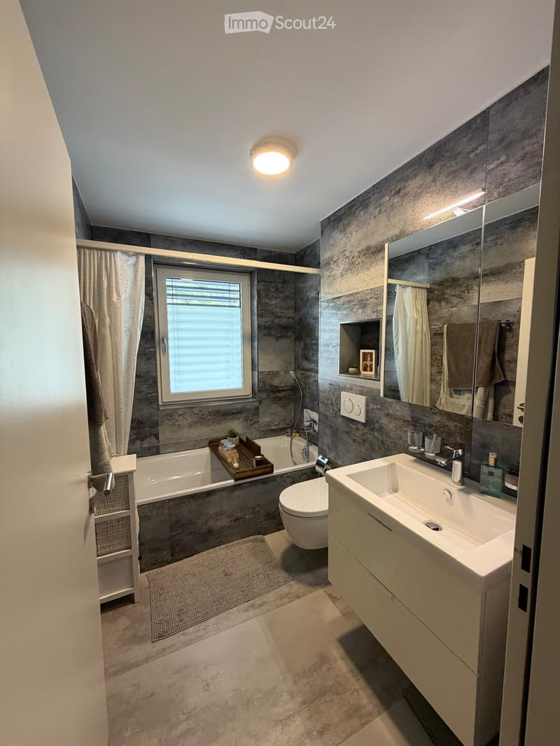 Modern bathroom with grey tiled walls, a bathtub, toilet, sink, large mirror, and a window with blinds. Includes a shelf with decorative items and a small plant on the edge of the bathtub.
