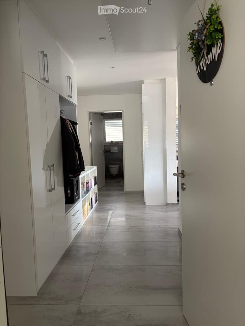 Modern hallway with white walls, grey tiled floor, built-in storage unit with books, coat rack, and a small bathroom visible at the end.