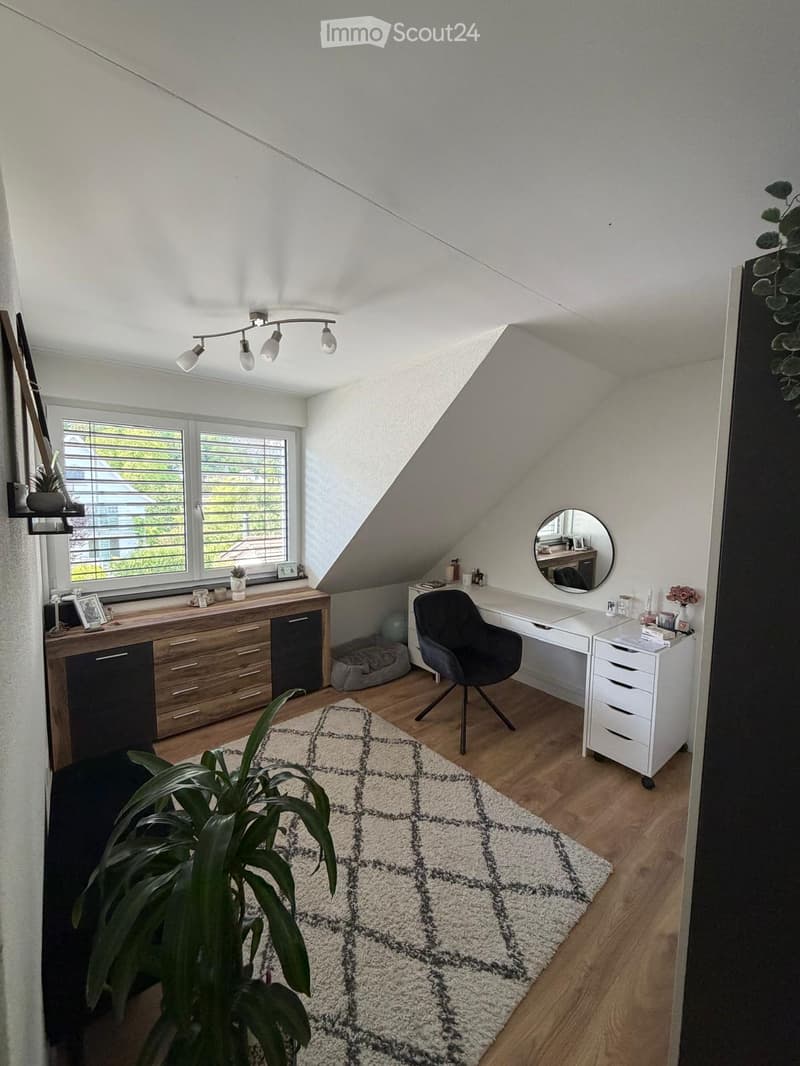 Bright attic room with wooden floors, large window, slanted ceiling, and modern furniture including a desk, chair, cabinet, and a round mirror.