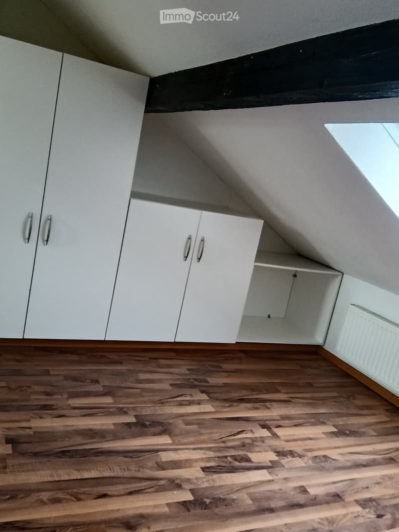 An attic space with wooden flooring and white built-in cabinets, featuring a slanted ceiling and a small window letting in natural light.