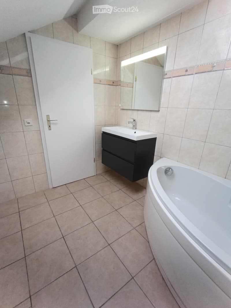 Modern bathroom with beige tiled walls and floor, white sink with black cabinet, bathtub, and illuminated mirror above the sink.