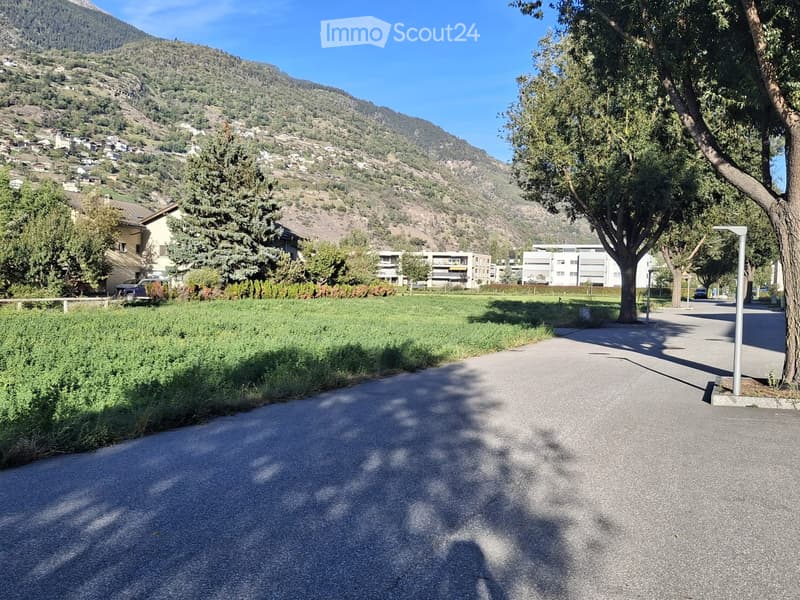 A paved road leads through a green area with trees and houses in the background, with a mountainous landscape visible in the distance.