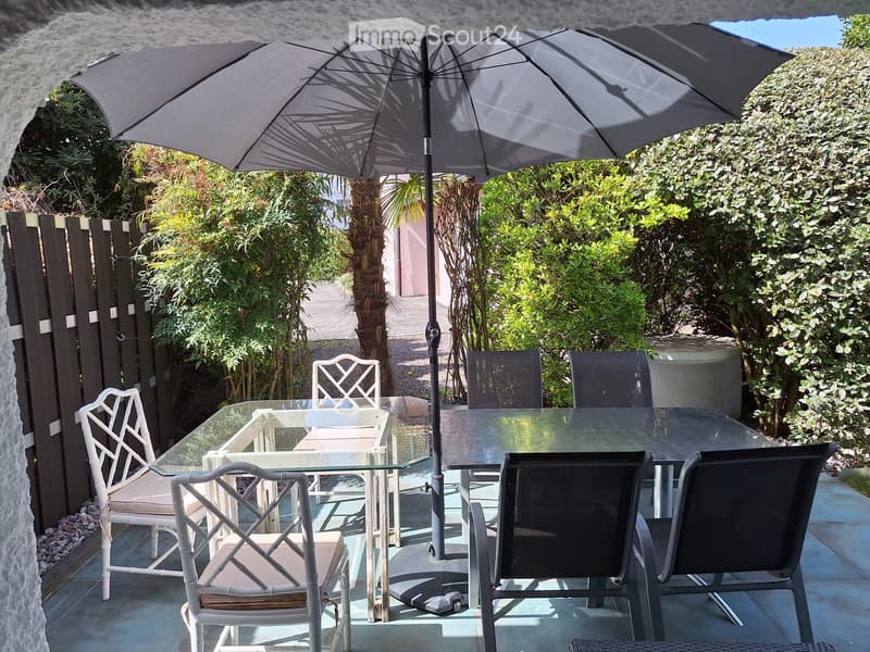 Outdoor patio with glass table, white and black chairs, large umbrella, surrounded by greenery and a stone wall.