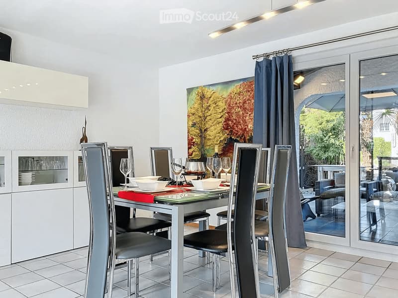 Modern dining room with a sleek glass dining table, six black leather chairs, wine glasses, bowls, and a modern painting on the wall. The room features white tiled flooring, white cabinets, and large glass doors leading to a balcony.