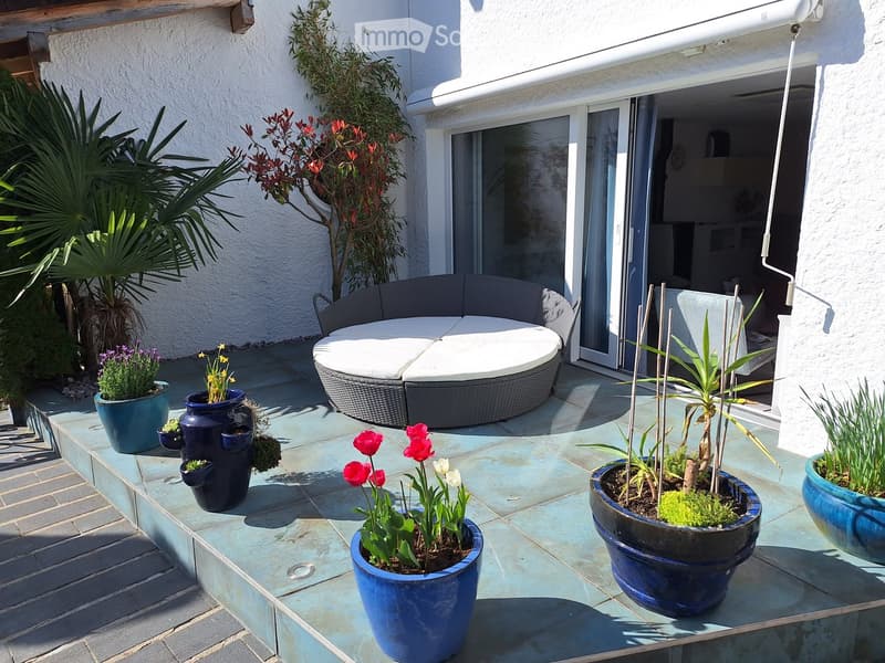 A modern outdoor patio with a round wicker sofa, multiple potted plants, and colorful flowers arranged on a tiled surface.