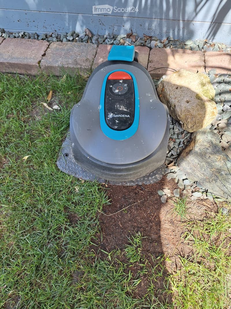 A robotic lawn mower is placed on a patch of grass near a stone border and a paved area.