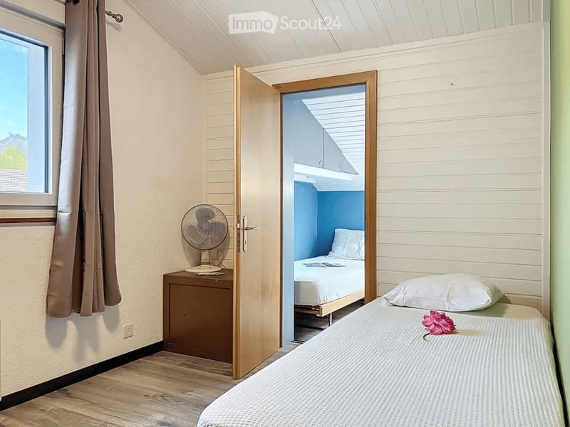 Cozy bedroom with a single bed, nightstand, and fan. The room features wooden flooring, white walls, and a slanted ceiling. An open door leads to another room with a bed and blue walls.