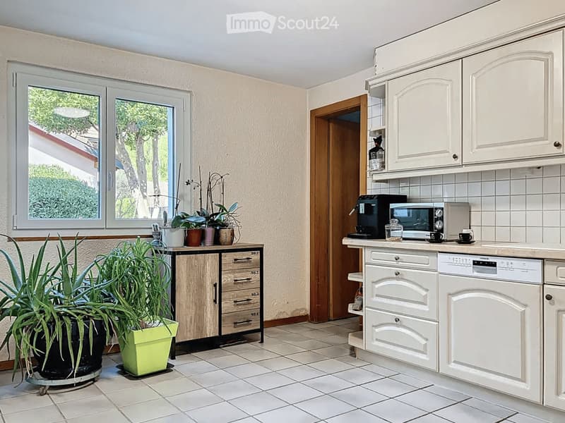Bright kitchen with white cabinets, tiled floor, and a window overlooking greenery. Includes a dishwasher, microwave, coffee maker, and a small table with potted plants.