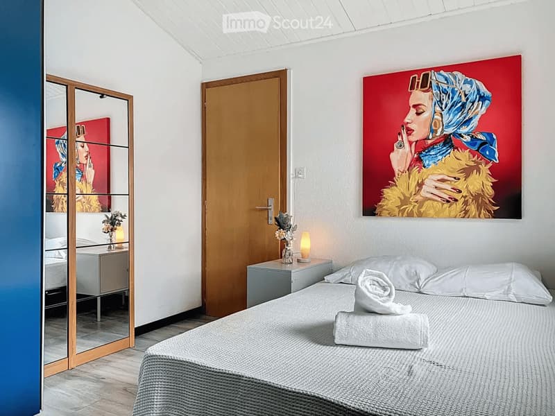 Modern bedroom with a grey bed, white pillows, nightstand, mirror, and artwork on the wall.