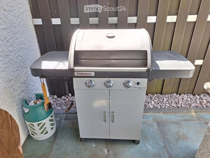 A modern gas grill with control knobs and a storage cabinet, placed on a tiled patio next to a gas tank and a brush.