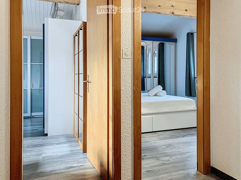 Modern bedroom with light wooden flooring, white walls, and a neatly made bed with pillows. The room features a large window with dark curtains and a built-in wardrobe with mirrored doors.