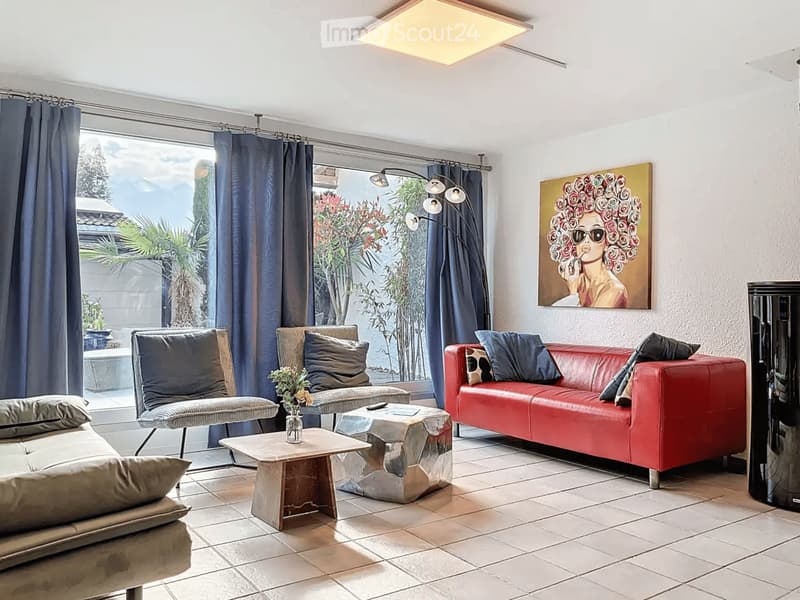 Modern living room with large windows offering a scenic view, featuring a red leather sofa, a grey armchair, a coffee table, and a floor lamp.