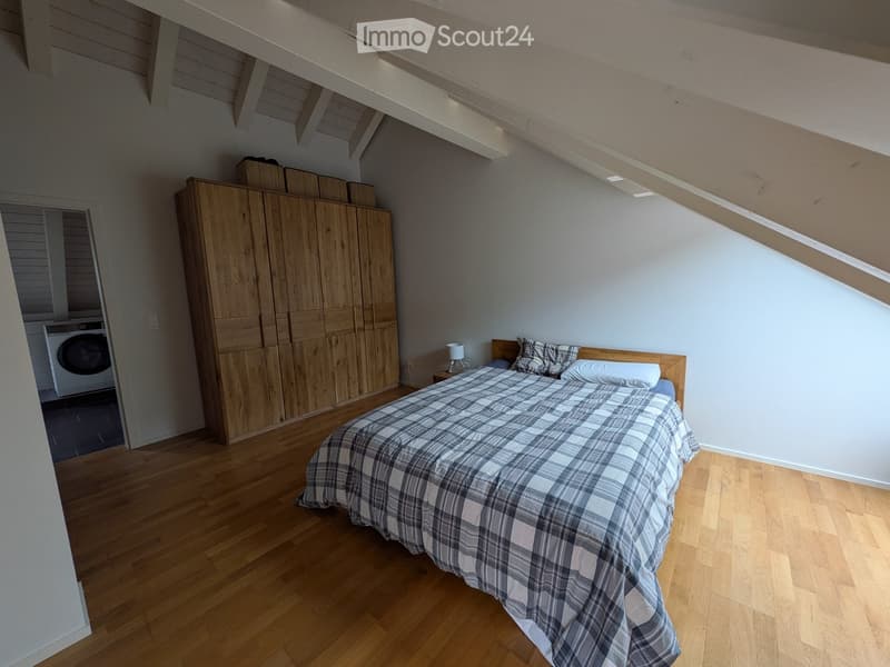 Bedroom with wooden floor, slanted ceiling, large wardrobe, bed with plaid duvet, nightstand with lamp, and visible washing machine in adjacent area.