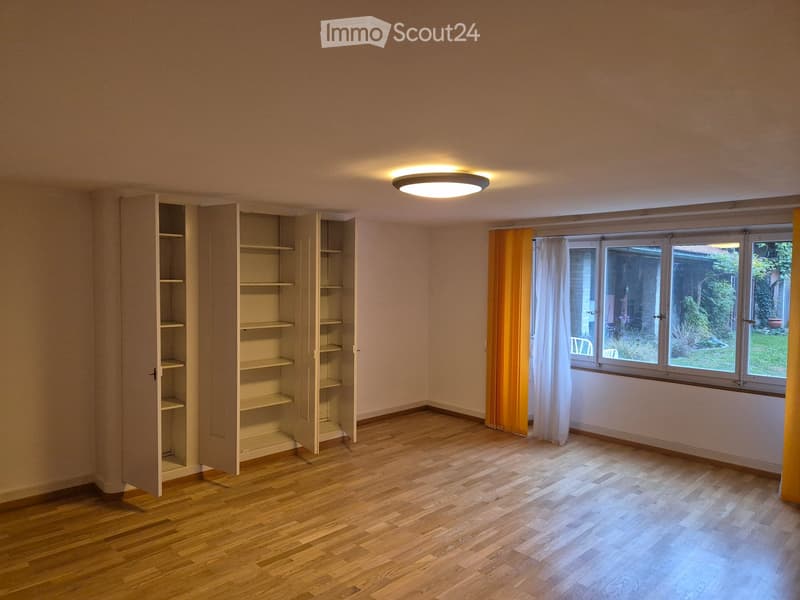 Empty room with wooden flooring, built-in shelves, large windows with curtains, and a ceiling light fixture.