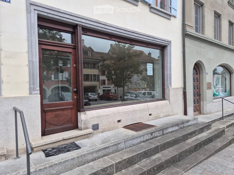 Entrance to a building with a wooden door, glass windows, and steps leading up to it. The building has a light-colored facade and is located on a street with other buildings visible in the background.