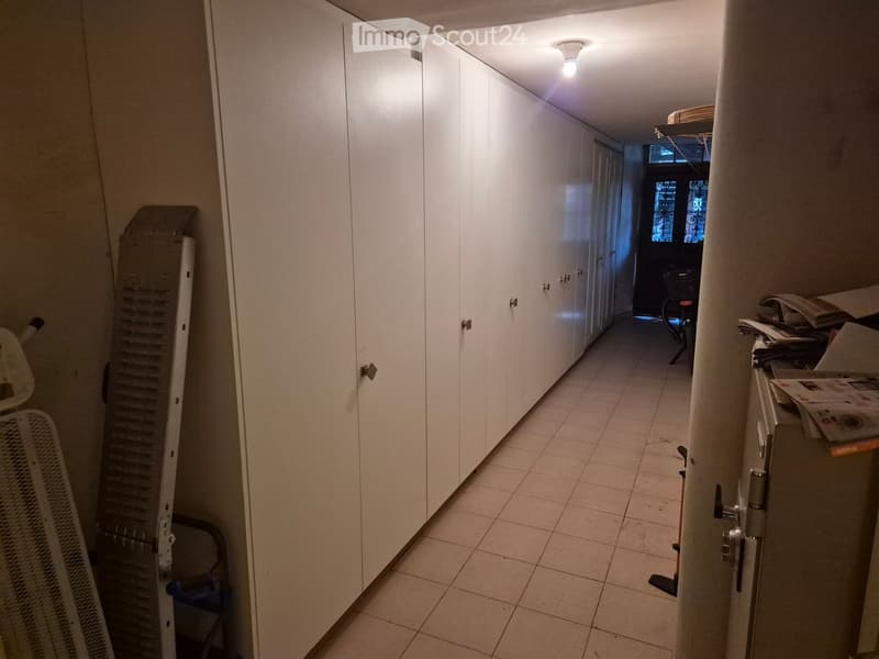 A narrow hallway with white tiled flooring, white walls, and built-in white cabinets on one side. The hallway has a ceiling light and leads to a room with a window and some furniture visible at the end.