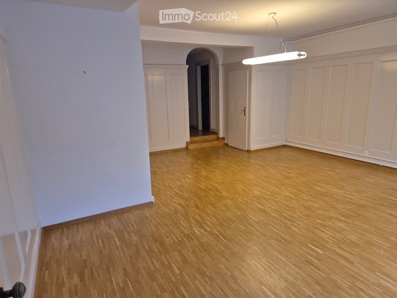 Empty room with wooden flooring, white walls, and a hanging light fixture. There are two doors and an archway leading to another space.