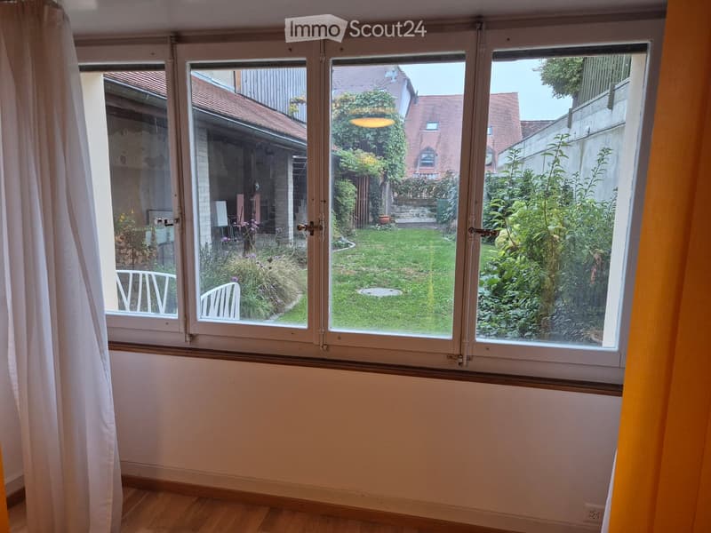 Empty room with wooden flooring, large window offering a view of a garden, and a curtain partially drawn to the left.