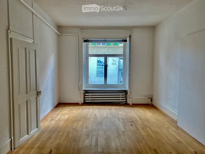 Empty room with wooden flooring, white walls, a single window with a radiator below, and a closed door on the left side.