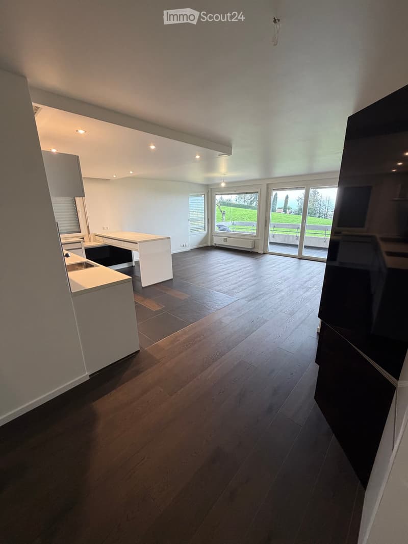 Open-plan unfurnished room with wooden flooring, white walls, and large windows leading to a balcony. Minimalist design with a small kitchen area visible on the left.