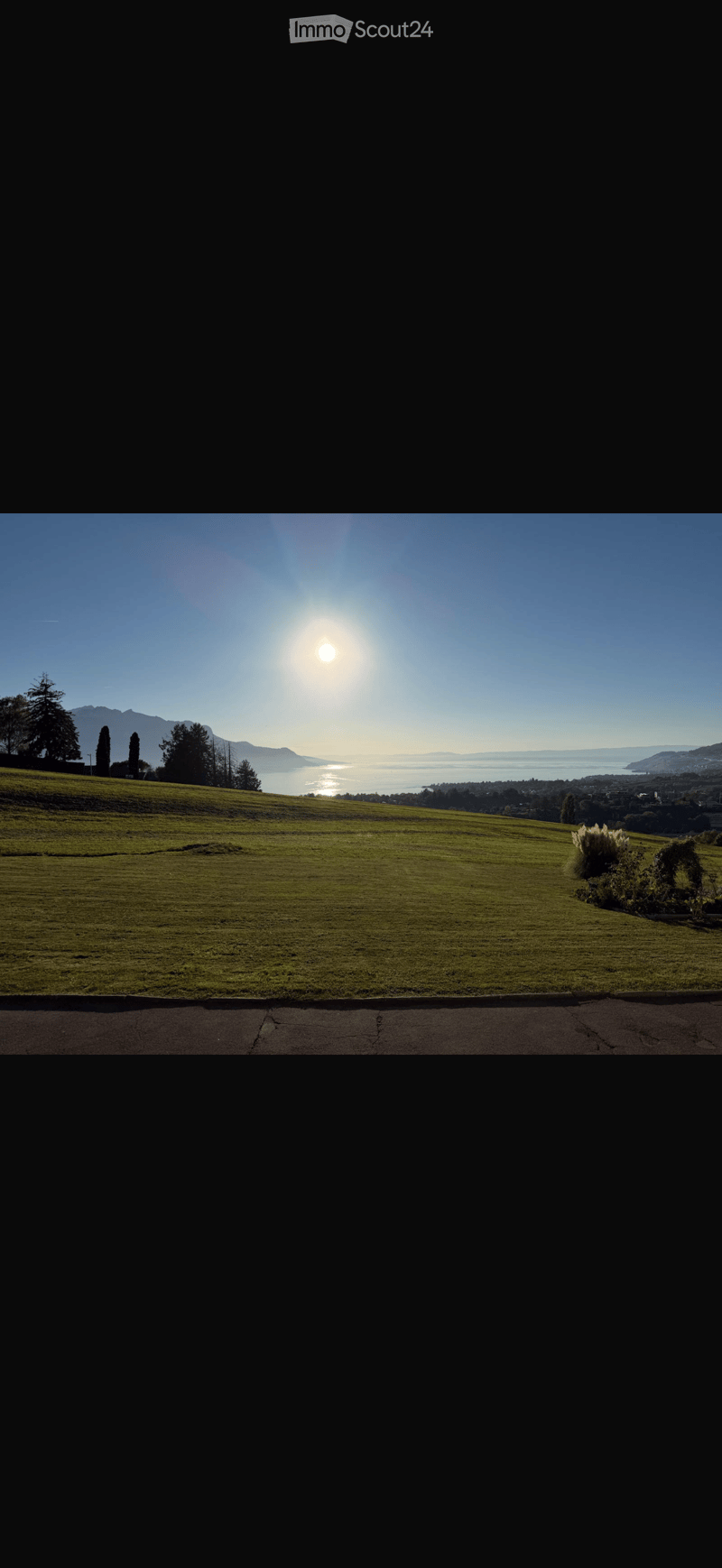 A wide, open grassy field with a clear view of a lake and distant mountains under a bright sun.