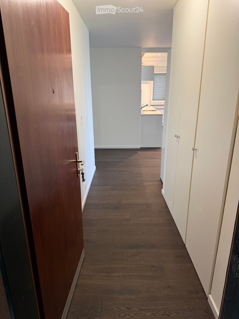 Empty hallway with wooden flooring, white walls, and a partially open door leading to a kitchen area.