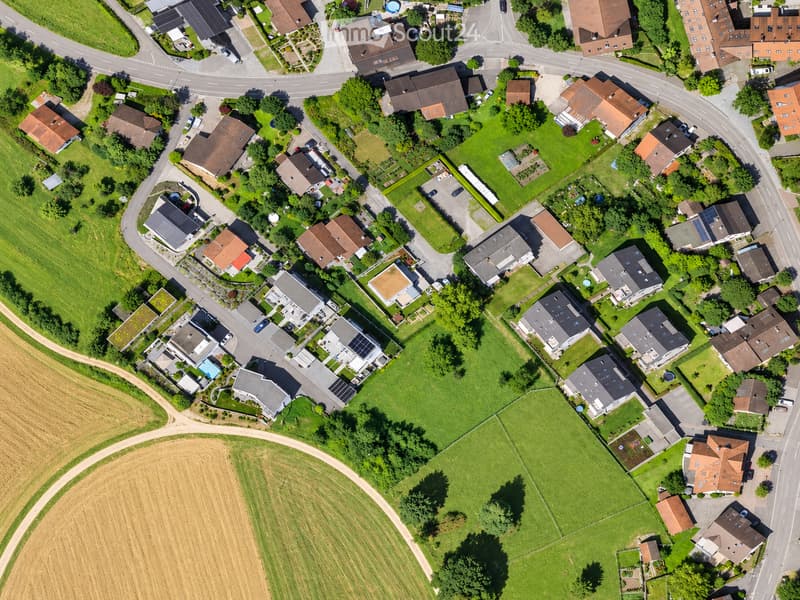 Aerial view of a suburban neighborhood with multiple houses, green lawns, and surrounding trees. Houses have varied roof styles and colors, with driveways and parked cars visible.
