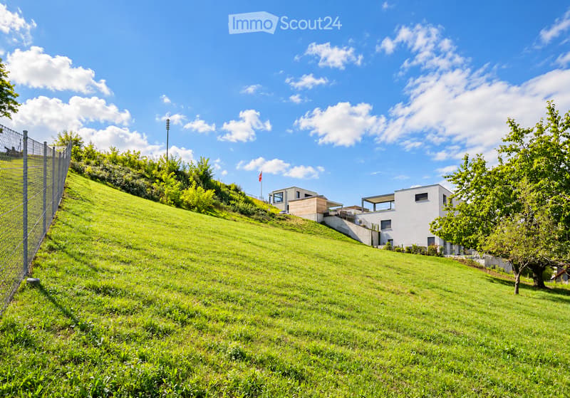 Modern two-story house on a grassy hillside with a fence and trees in the background.