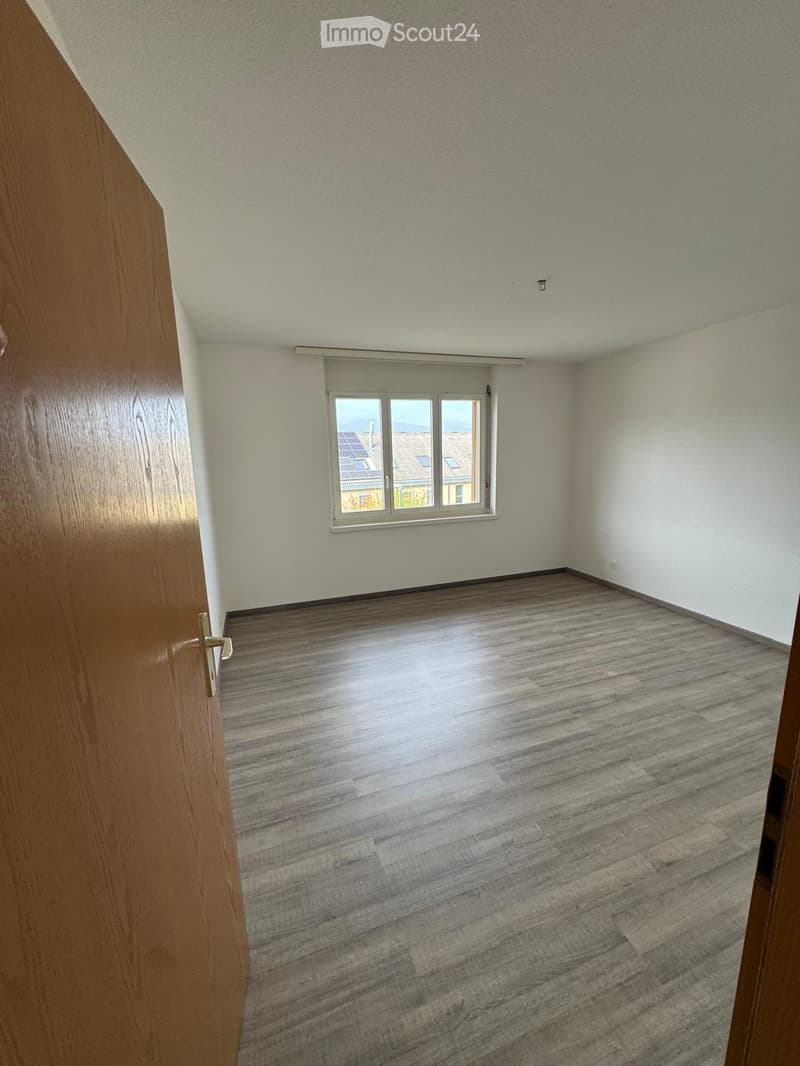 Empty room with wooden flooring, white walls, and a window showing a view of rooftops.