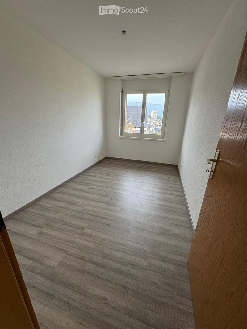 Empty room with wooden flooring, white walls, and a window offering a view of the city and mountains.