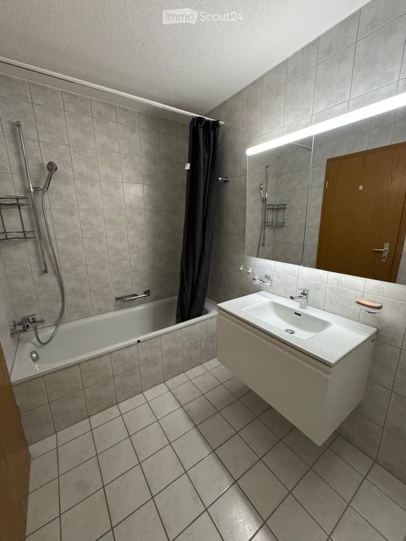 Modern bathroom with a bathtub, sink, mirror, and shower area with a curtain. Grey tiles and a wooden cabinet are visible.
