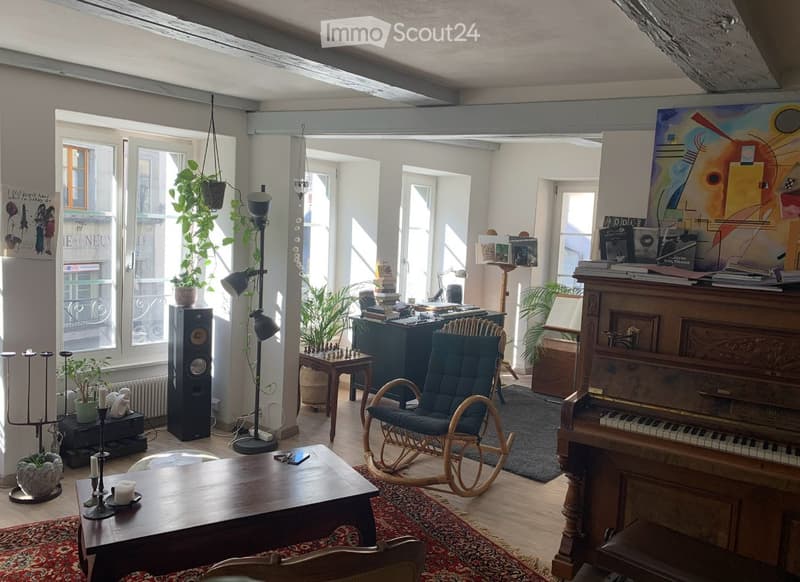 Bright, airy room with wooden floors, large windows, and modern furnishings including a piano, a rocking chair, a coffee table, and a speaker system.