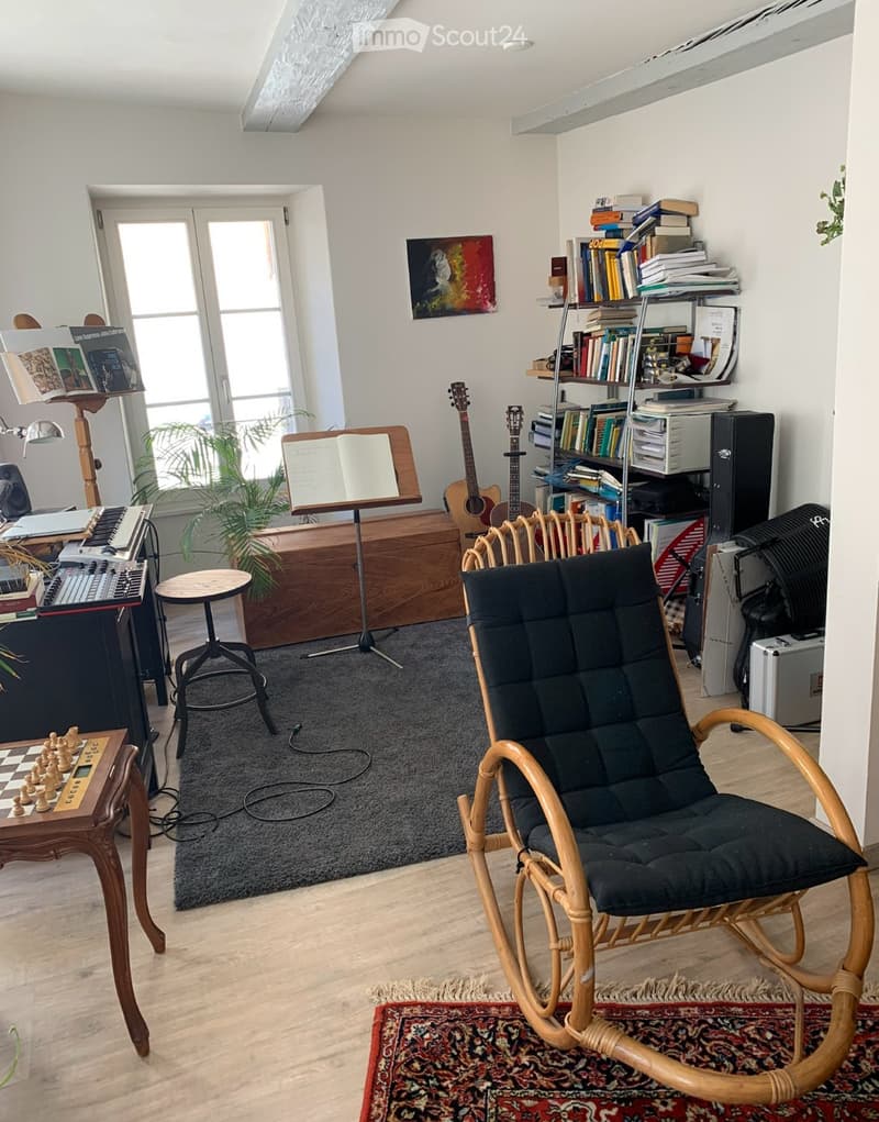 A well-lit room with a wooden floor, a large window, a black rocking chair, a small table with a chessboard, a bookshelf filled with books, a guitar, a keyboard, and a plant near the window.