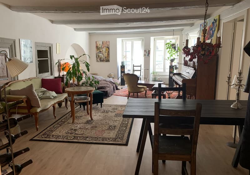 A spacious, well-lit living and dining area with wooden floors, a patterned rug, a sofa, a dining table, chairs, a piano, and decorative lamps.