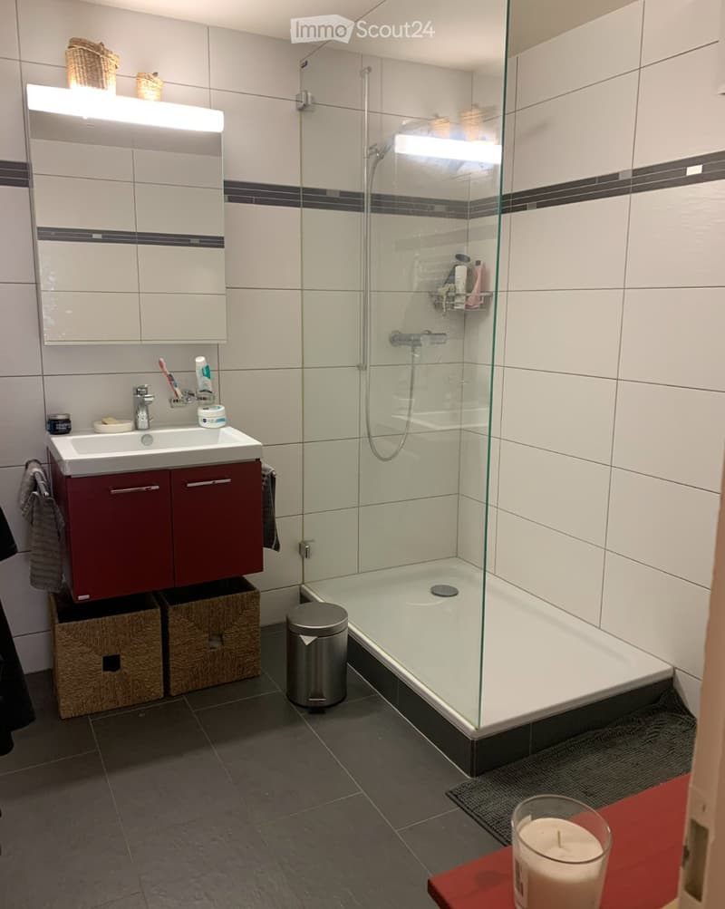 Modern bathroom with white tiles, dark grout borders, a red cabinet under the sink, a glass-enclosed shower, and wicker baskets on the floor.