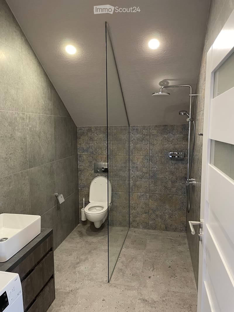 Modern bathroom with a toilet, sink, and separate shower area. Features grey tiles, a glass partition, and built-in storage.
